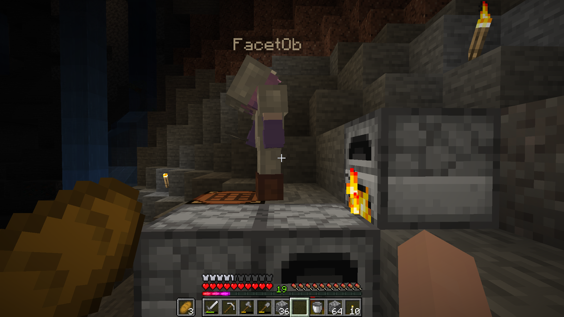 Alice verwendet eine Werkbank. Daneben stehen drei Öfen. Links sind Wasserfälle in einem dunkleren Teil der Höhle zu sehen.