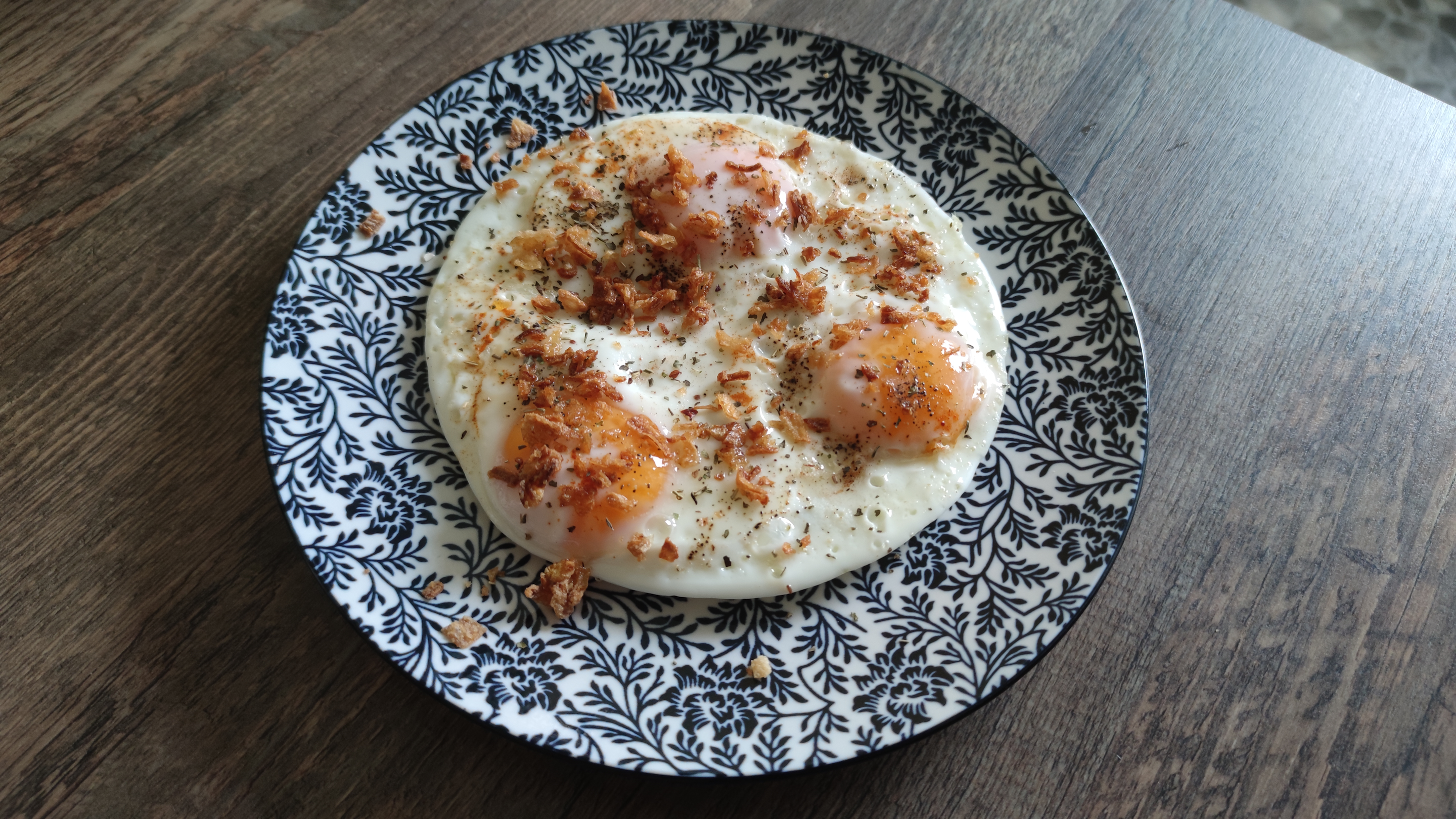 Auf einem blau‐weißen Teller liegt eine runde Masse aus drei gebratenen Eiern, übersät mit verschiedenen Gewürzen und Röstzwiebeln.